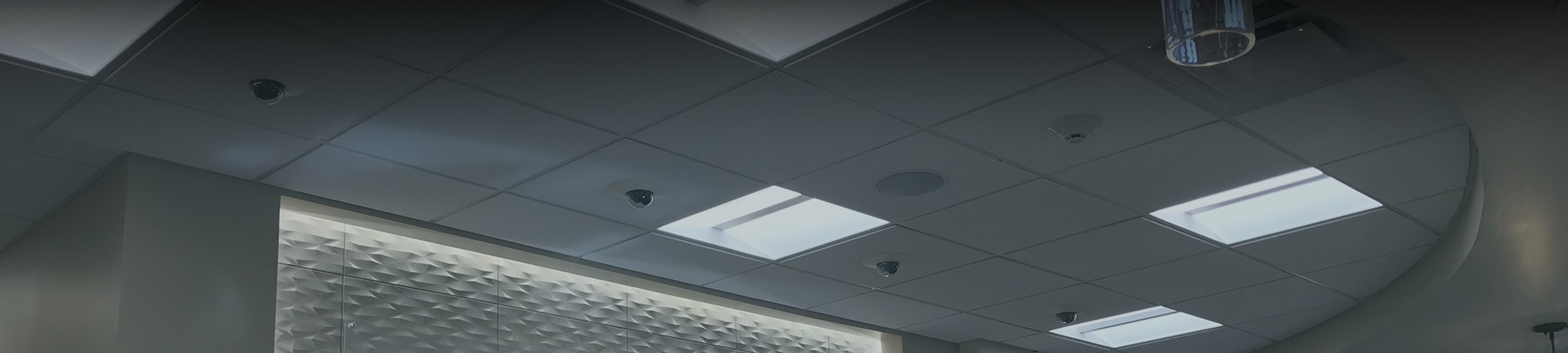 Commercial acoustical tile ceiling with integrated security cameras, lights, sprinkler heads, and speakers in a medical facility