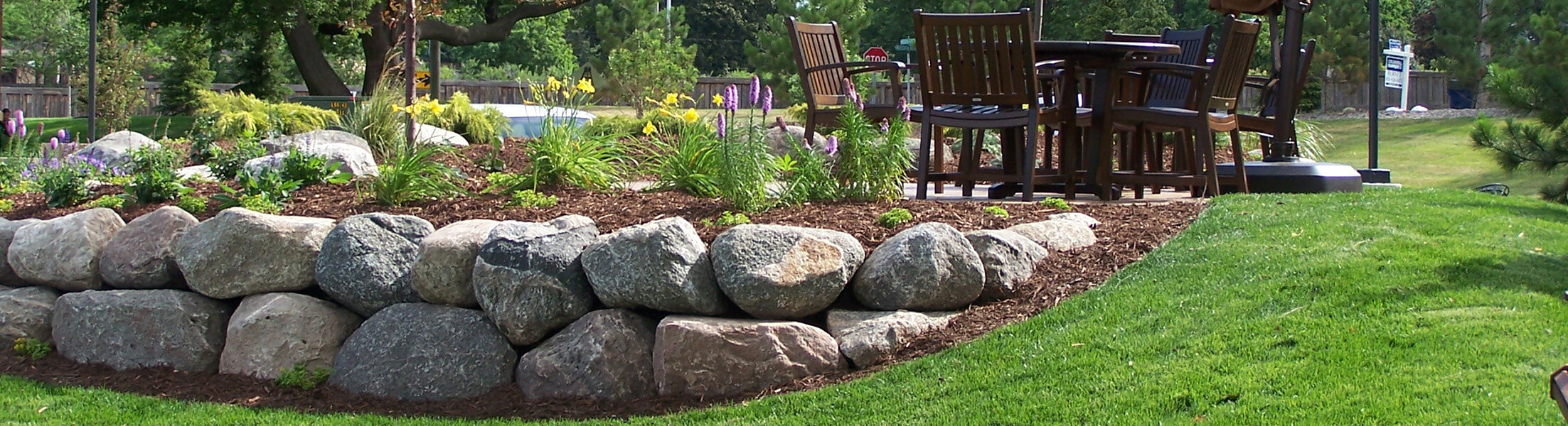 Flowering plant garden surrounded by fieldstone boulders next to a backyard patio